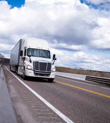 Classic modern new white big rig semi truck with a trailer moving on a smooth cloudy highway transporting a cargo of industrial products for livelihood all across America